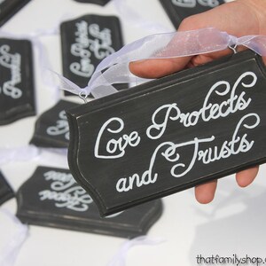 May include: Black wooden sign with white lettering that reads "Love protects and trusts". The sign is held by a hand and has a white ribbon attached.