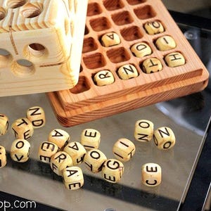 May include: A wooden word game set featuring a light-colored storage box with circular holes, a grid-patterned wooden board containing lettered cubes, and several loose wooden cubes with black printed letters scattered on the surface.