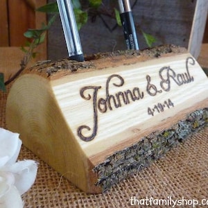 Puede incluir: Un portaplumas de madera rústico con los nombres "Johanna & Paul" y la fecha "4-19-14" grabados en él. El portaplumas está hecho de una rama de árbol natural con corteza aún adherida.
