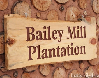 Puede incluir: Un letrero de madera con las palabras "Bailey Mill Plantation" talladas en él en letras marrones. El letrero está colgado en una pared hecha de troncos apilados.