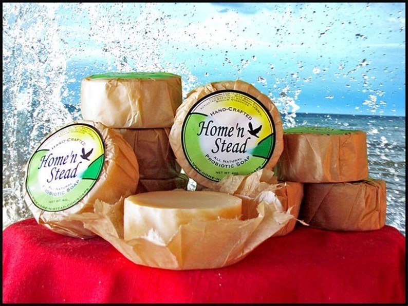 May include: A stack of round, brown, paper-wrapped bars of soap with a green and yellow label that reads "Hand-Crafted Home'n Stead All Natural Probiotic Soap Net Wt 4oz".