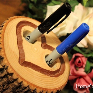Puede incluir: Un portaplumas de madera de tronco con un diseño en forma de corazón tallado en la parte superior. Dos marcadores, uno negro y otro azul, se insertan en el portaplumas.