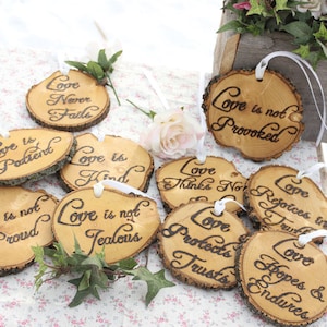 Puede incluir: Un conjunto de 10 adornos de madera con diferentes frases sobre el amor, cada uno con una cinta blanca. Las frases incluyen "El amor es paciente", "El amor es amable", "El amor nunca falla", "El amor no es orgulloso", "El amor no es envidioso", "El amor no es arrogante", "El amor se regocija en la verdad", "El amor espera y persevera", "El amor no piensa mal", y "El amor protege, confía".