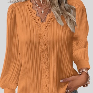 May include: An orange blouse with long, pleated sleeves and a V-neckline. The neckline features delicate lace trim. The blouse has a relaxed fit and is made of a lightweight fabric. The sleeves are gathered at the cuffs.