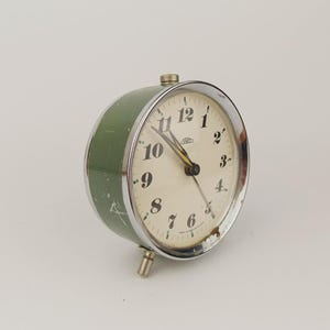 Könnte beinhalten: Vintage grüner und verchromter Wecker mit cremefarbenem Zifferblatt. Die Uhr hat schwarze Zahlen, eine Chromblende und einen kleinen Fuß. Auf dem Zifferblatt steht das Wort "Prim".