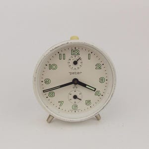 Puede incluir: Un despertador blanco vintage con una esfera color crema, manecillas negras y números verdes. El reloj tiene una pequeña perilla amarilla en la parte superior y tres patas plateadas. La palabra "peter" está impresa en la esfera.