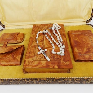May include: An antique religious set in a decorative box. The set includes a book, a rosary with a cross, a small wallet, and a larger wallet. The items are a warm brown colour and rest on a yellow surface.