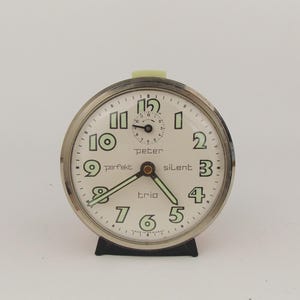 Puede incluir: Un reloj despertador plateado vintage con esfera crema. Tiene números negros, manecillas verdes brillantes y las palabras "peter", "perfekt", "silent" y "trio". El reloj tiene una base negra y una parte superior verde claro. La esfera mide unos 10 cm de diámetro.