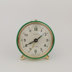 May include: A vintage green and gold alarm clock with a cream-coloured face. The clock face features black numerals and hands, with the text "ROSTOV" and "MADE IN USSR". The clock is approximately 7.6 cm in diameter.