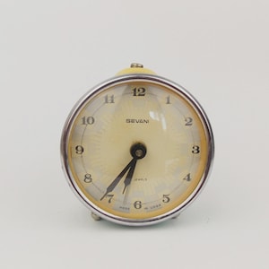 Peut inclure: Un réveil vintage avec un cadran crème et un cadre en métal argenté. Le cadran de l'horloge comporte des chiffres et des aiguilles noirs, avec la marque "SEVANI" visible. L'horloge est ronde et a un dessus jaune.