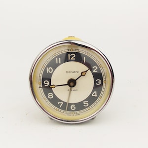 Può includere: Una sveglia rotonda vintage con quadrante color crema e numeri neri. L'orologio ha una lunetta color argento e una base gialla. Il quadrante dell'orologio indica "SEVANI" e "4 JEWELS".