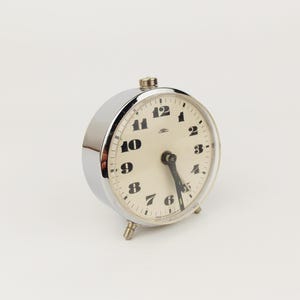 Könnte beinhalten: Ein Vintage-Wecker aus Chrom mit cremefarbenem Zifferblatt und schwarzen Ziffern. Die Uhr hat eine runde Form, zwei schwarze Zeiger und drei kleine Füße. Auf dem Zifferblatt steht der Markenname "Laco".