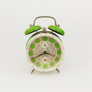 Könnte beinhalten: Vintage grün-weißer Wecker mit dekorativem Zifferblatt. Die Uhr hat grüne Glocken, einen silbernen Griff und grüne römische Ziffern. Das Zifferblatt hat ein florales Design und zwei kleine Zifferblätter. Ein klassisches Retro-Design.