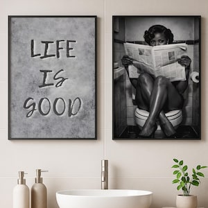 May include: Two framed wall art prints in a bathroom. The left print features a gray textured background with the black text "LIFE IS GOOD". The right print is a black and white photograph of a person sitting on a toilet reading a newspaper.