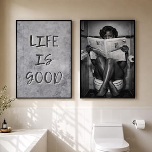 May include: Two framed wall art prints hanging above a bathroom vanity. The left print features a gray textured background with the black text "LIFE IS GOOD". The right print is a black and white photograph of a person reading a newspaper on a toilet.