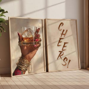 May include: Two framed art prints. One features a hand adorned with gold bracelets and rings, holding a glass of amber liquid with a large ice cube. The other print says "CHEERS!" in brown script.