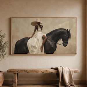 Può includere: Un'opera d'arte incorniciata raffigura una persona con un abito bianco e un cappello a tesa larga a cavallo di un cavallo nero. L'opera è esposta sopra una panca di legno con una coperta beige. La stanza ha una palette di colori neutri.