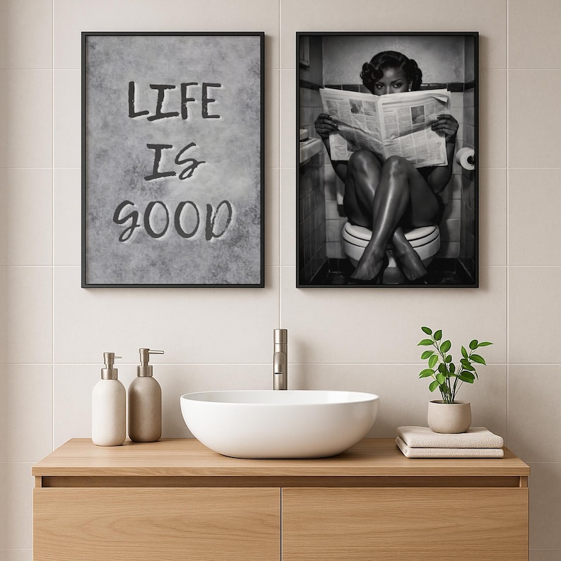 May include: Two framed wall art prints in a bathroom. The left print features a gray textured background with the black text "LIFE IS GOOD". The right print is a black and white photograph of a person sitting on a toilet reading a newspaper.