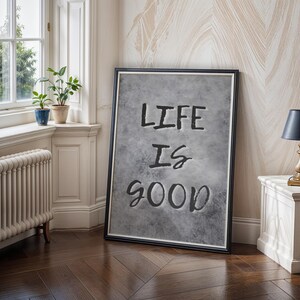 May include: A rectangular framed wall art piece featuring a textured gray background with the black text "LIFE IS GOOD" centered in the middle. The artwork is enclosed in a thin black frame with a white inner border, displaying a clean design.