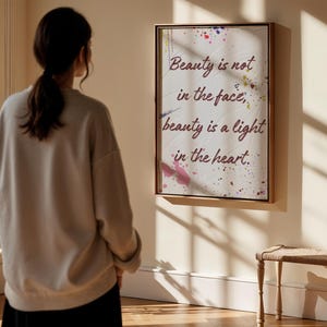 May include: A framed print with the quote "Beauty is not in the face, beauty is a light in the heart." The artwork features a white background with colourful paint splatters. It is displayed on a wall. A person is standing in front of the artwork.