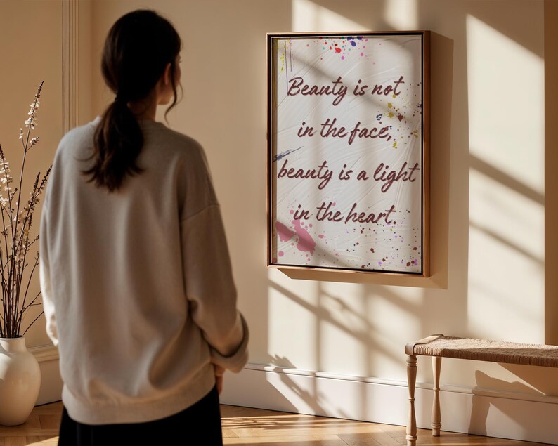 May include: A framed print with the quote "Beauty is not in the face, beauty is a light in the heart." The artwork features a white background with colourful paint splatters. It is displayed on a wall. A person is standing in front of the artwork.