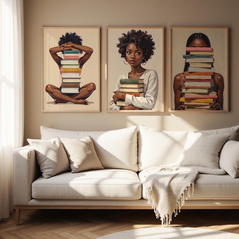 May include: Three framed art prints featuring illustrations of Black women with books. One print shows a woman sitting with a stack of books, another shows a woman holding books, and the third shows a woman with books covering her face. The prints are displayed above a white sofa.