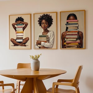 May include: Three framed art prints featuring portraits of Black women with stacks of books. The women are holding or interacting with the books. The prints are displayed above a wooden table and chairs.