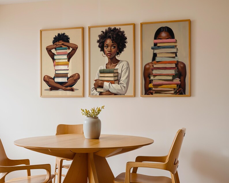 May include: Three framed art prints featuring portraits of Black women with stacks of books. The women are holding or interacting with the books. The prints are displayed above a wooden table and chairs.