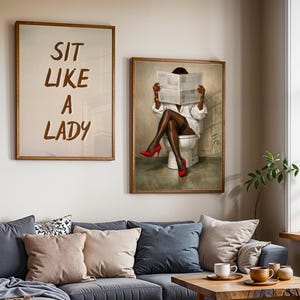 May include: Two framed art prints hang above a gray sofa. One print reads "SIT LIKE A LADY" in brown letters. The other depicts a person in a white shirt and red heels reading a newspaper while seated on a toilet.
