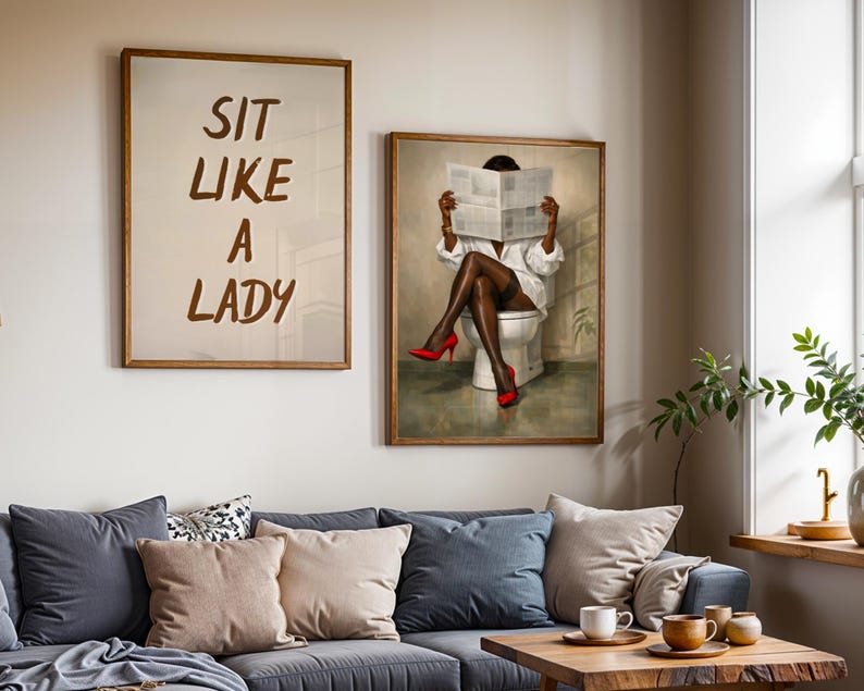 May include: Two framed art prints hang above a gray sofa. One print reads "SIT LIKE A LADY" in brown letters. The other depicts a person in a white shirt and red heels reading a newspaper while seated on a toilet.