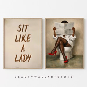 May include: Two framed art prints. One features the text "SIT LIKE A LADY" in brown lettering on a beige background. The other depicts a person in a white shirt, black stockings, and red heels, reading a newspaper while seated on a toilet.
