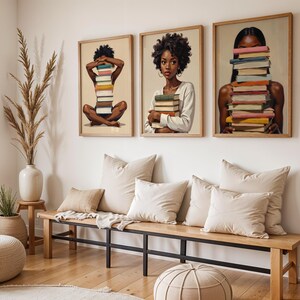 May include: Three framed art prints featuring illustrations of people with stacks of books. The artwork has a neutral color palette with wooden frames. The room also includes a wooden bench with pillows, a vase with tall grass, and a round ottoman.