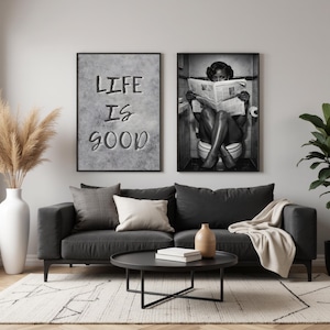May include: Two framed black and white wall art prints hanging above a dark gray sofa. The left print features the text LIFE IS GOOD in a bold font on a textured gray background. The right print shows a person sitting on a toilet reading a newspaper.