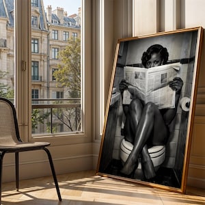May include: A large framed black and white wall art print depicting a person sitting on a toilet while reading a newspaper. The person has dark hair styled in waves and is positioned in a tiled bathroom setting. The artwork is encased in a gold frame.