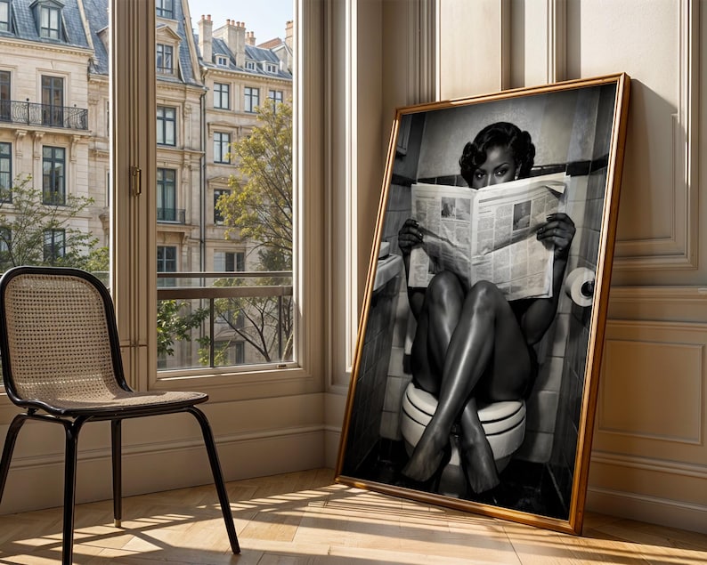 May include: A large framed black and white wall art print depicting a person sitting on a toilet while reading a newspaper. The person has dark hair styled in waves and is positioned in a tiled bathroom setting. The artwork is encased in a gold frame.