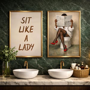 May include: Two framed art prints above white sinks. One print reads "SIT LIKE A LADY" in brown text. The other depicts a person in a white shirt and red heels reading a newspaper while seated on a toilet. Gold frames.