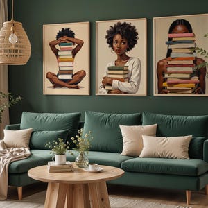 May include: Three framed art prints featuring Black women holding stacks of books. The artwork has a warm color palette and is displayed above a green velvet sofa with beige pillows. A wooden coffee table and decorative plants complete the room's decor.