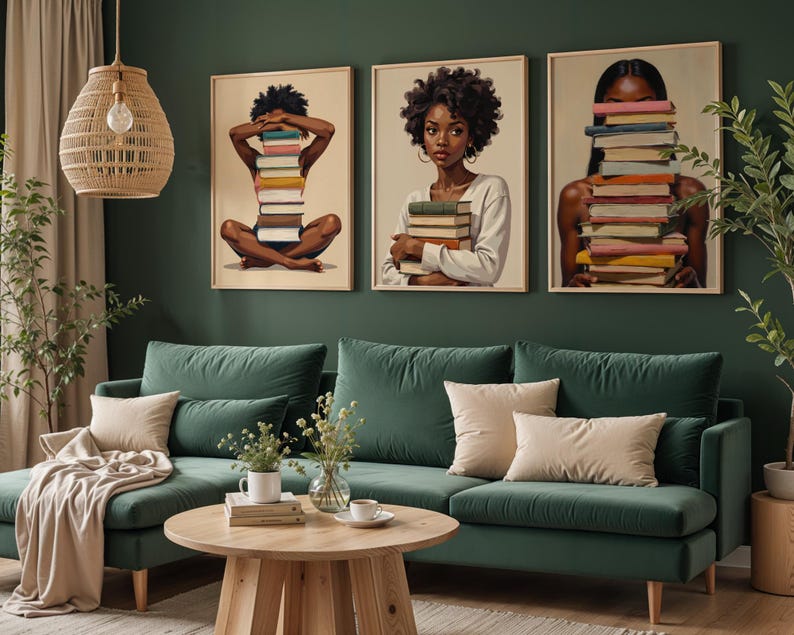 May include: Three framed art prints featuring Black women holding stacks of books. The artwork has a warm color palette and is displayed above a green velvet sofa with beige pillows. A wooden coffee table and decorative plants complete the room's decor.