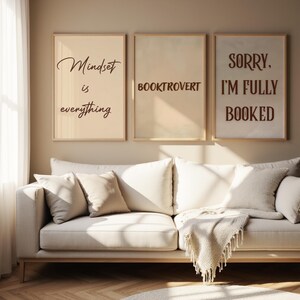 May include: Three framed prints with text: "Mindset is everything", "BOOKTROVERT", and "SORRY, I'M FULLY BOOKED". The prints are in neutral tones and hang above a cream sofa with cushions and a fringed throw. The room has a neutral colour scheme.