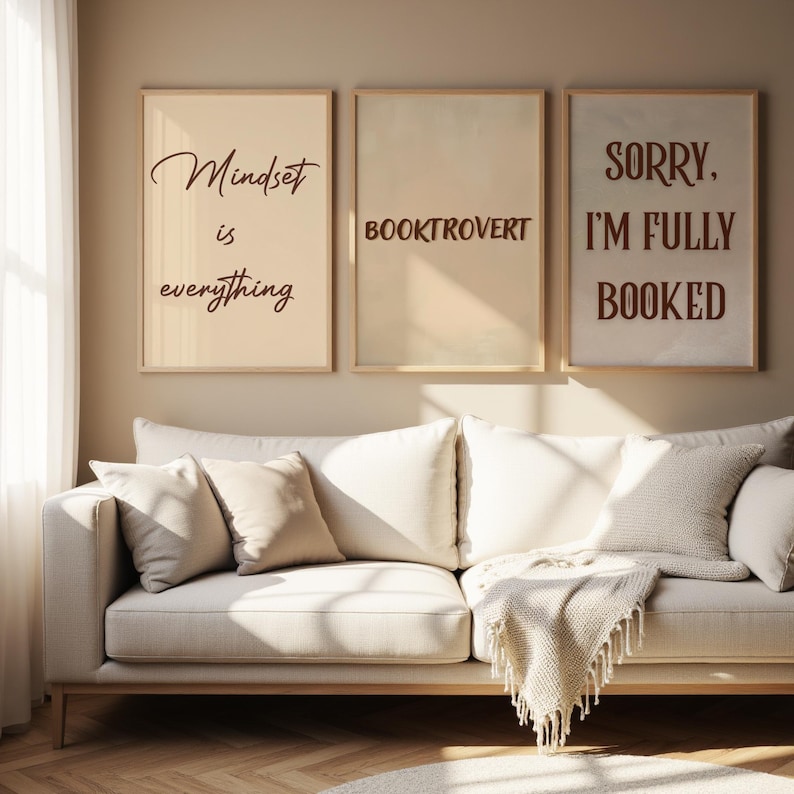 May include: Three framed prints with text: "Mindset is everything", "BOOKTROVERT", and "SORRY, I'M FULLY BOOKED". The prints are in neutral tones and hang above a cream sofa with cushions and a fringed throw. The room has a neutral colour scheme.