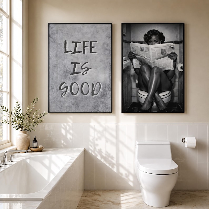 May include: Two framed wall art prints hanging above a bathroom vanity. The left print features a gray textured background with the black text "LIFE IS GOOD". The right print is a black and white photograph of a person reading a newspaper on a toilet.