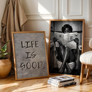 May include: Two framed wall art prints. One print features a gray textured background with the black text "LIFE IS GOOD". The second print is a black and white photograph of a person sitting on a toilet reading a newspaper in a tiled bathroom.
