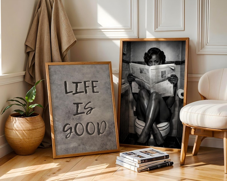 May include: Two framed wall art prints. One print features a gray textured background with the black text "LIFE IS GOOD". The second print is a black and white photograph of a person sitting on a toilet reading a newspaper in a tiled bathroom.