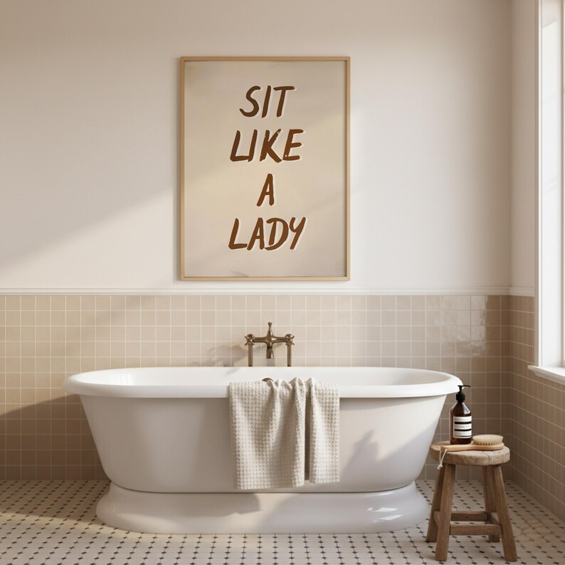 May include: A bathroom scene featuring a white, freestanding bathtub with a towel draped over the edge. Above the tub, a framed print reads "SIT LIKE A LADY" in brown lettering. A small wooden stool holds a bottle and brush.
