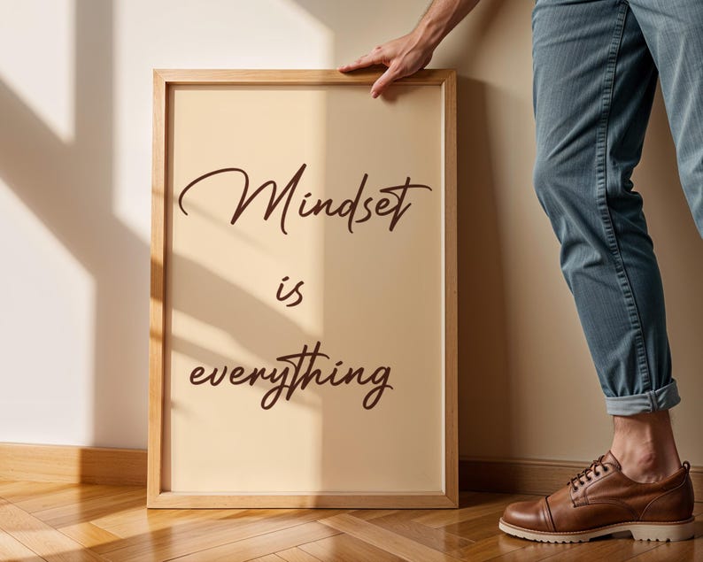 May include: A framed print with the words "Mindset is everything" in a stylish, cursive font. The frame is a light wood color, and the background is a soft beige. The print is leaning against a wall.