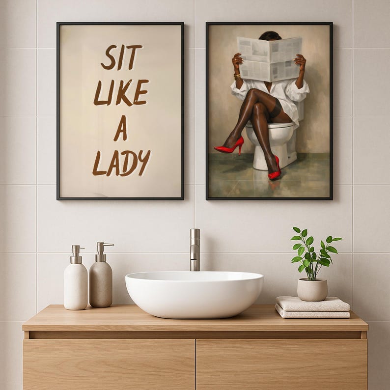 May include: Two framed art prints. One reads "SIT LIKE A LADY" in brown script. The other depicts a person in a white shirt and red heels reading a newspaper while seated on a toilet. The art is displayed above a wooden vanity with a white sink.