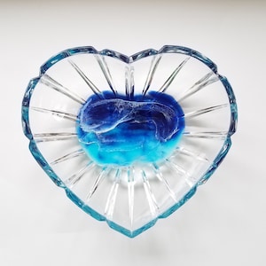 Può includere: Un piatto di vetro a forma di cuore con un'infusione di resina blu e turchese all'interno. La resina ha un motivo ondulato.