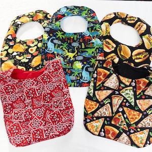 May include: Five different baby bibs with various patterns. One bib has a red and white bandana pattern, one has a black and green pizza pattern, one has a black and yellow hamburger pattern, one has a blue and green dinosaur pattern, and one has a black and green avocado pattern.