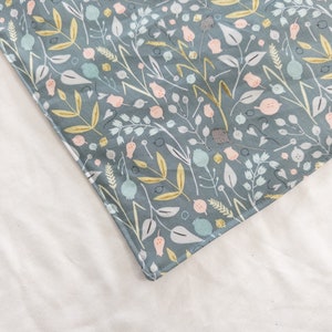 May include: A corner of a fabric item with a botanical pattern. The design features a teal background with white, yellow, and pink floral and leaf elements. The item is placed on a white surface.