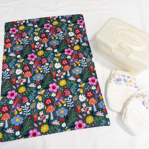 May include: A green fabric changing mat with a floral and mushroom pattern.  The mat is next to a white wipes container with the word "Huggies" on it.  Two white baby diapers with colorful designs are also in the image.